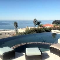 Infinity pool overlooking the ocean, with lounge seating. Blue water, sunny sky.
