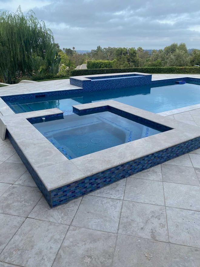 Swimming pool and spa with blue tile and light stone surround. Cloudy day, outdoor setting.