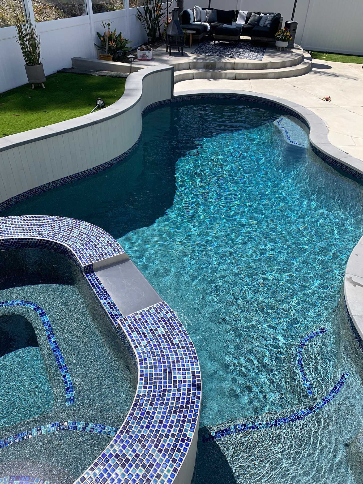 Swimming pool with blue tiles and clear water, surrounded by concrete and greenery.