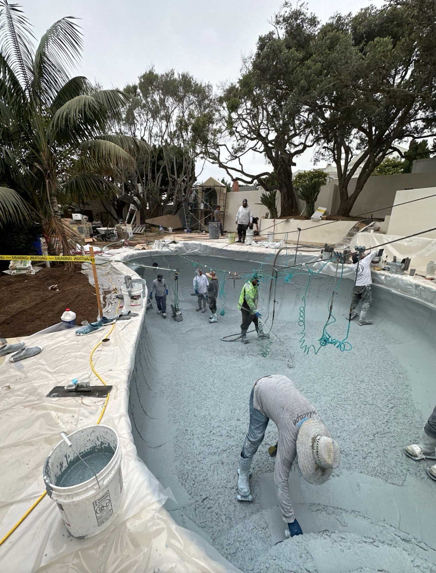 Construction workers applying plaster to a swimming pool. Several people are inside the pool, with tools and materials. Outdoors, cloudy day.