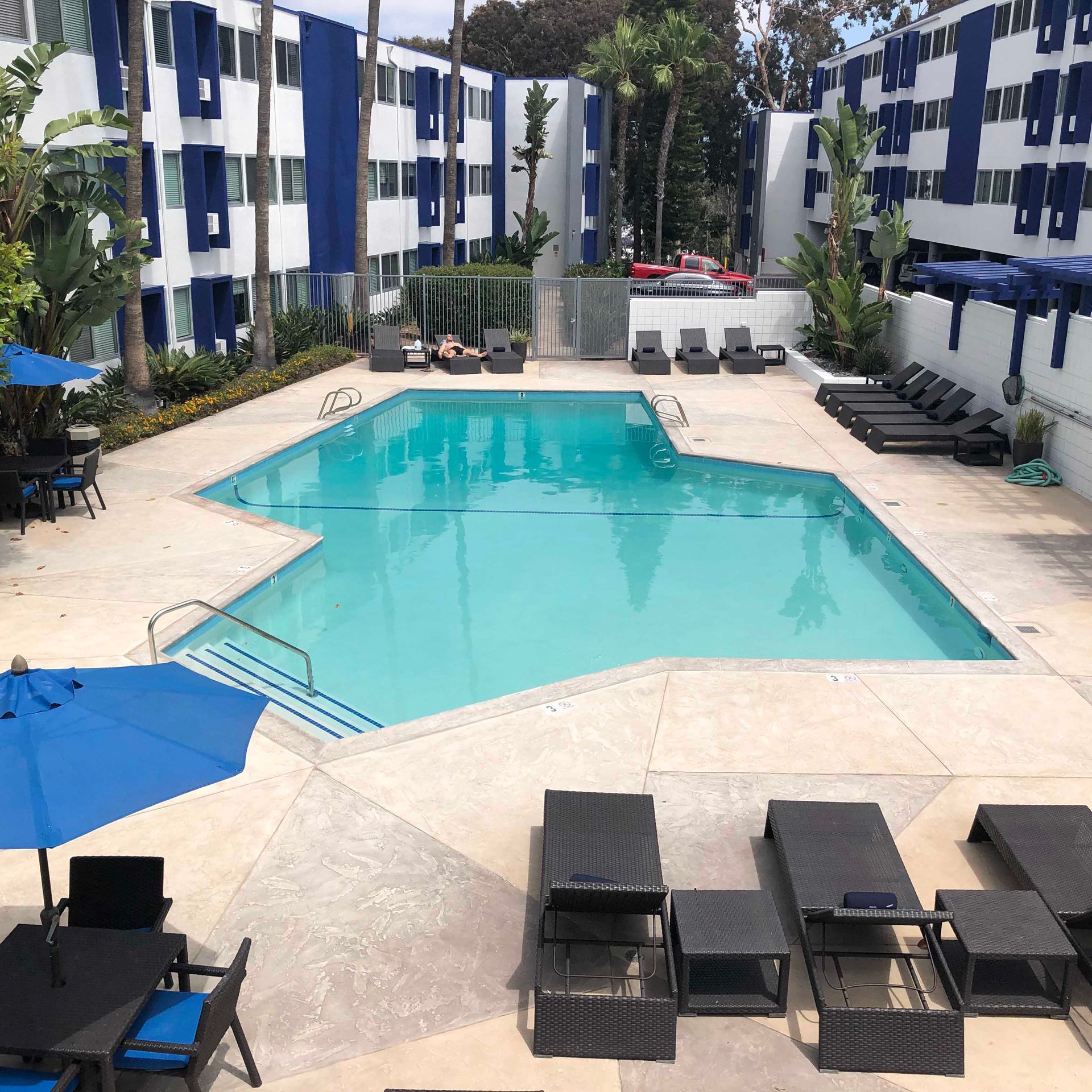Swimming pool surrounded by buildings with blue and white accents; lounge chairs, tables, and umbrellas.