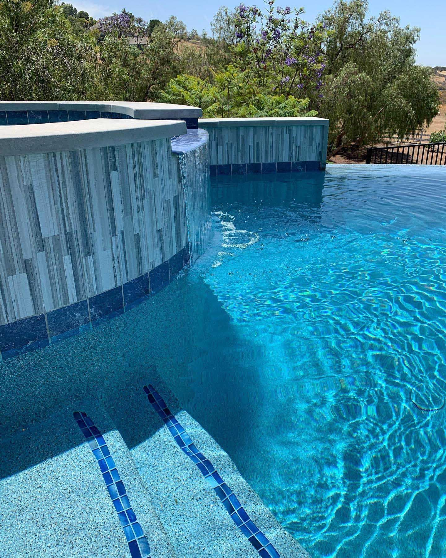 Blue tiled infinity pool with clear water, steps, and surrounding greenery under a sunny sky.