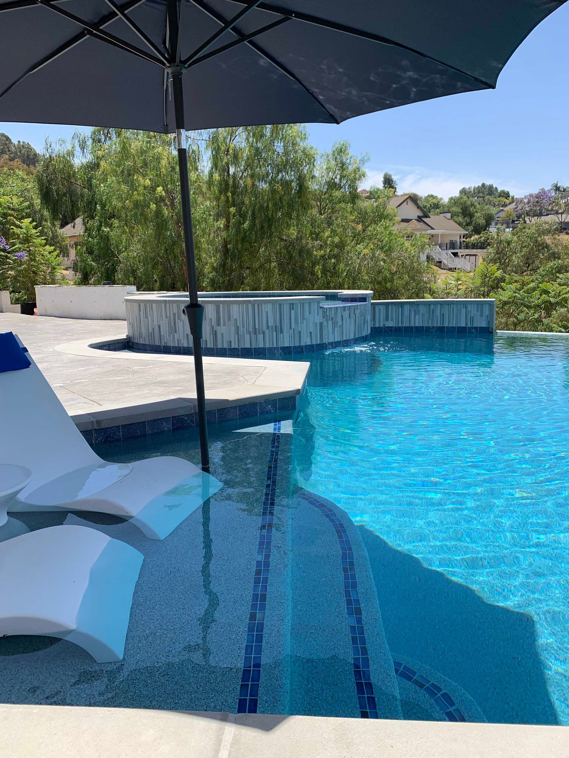 A blue pool with steps, a black umbrella, and white lounge chair, surrounded by greenery and a bright blue sky.