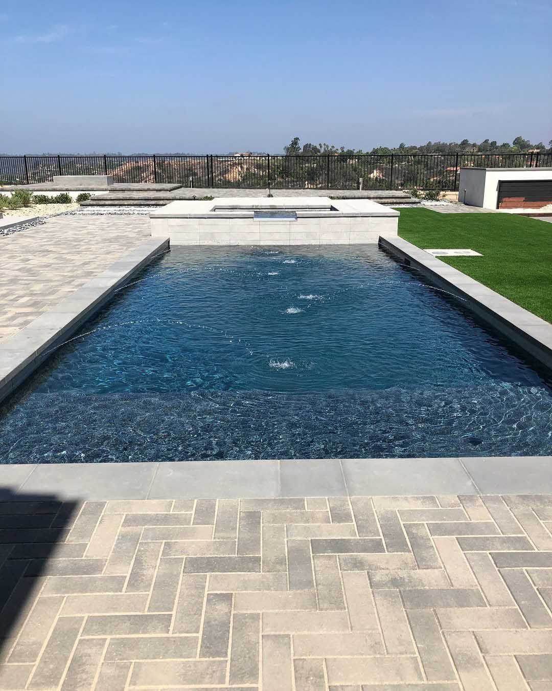 Rectangular pool with water fountains, surrounded by grey brick patio and green lawn. Distant view of landscape and blue sky.