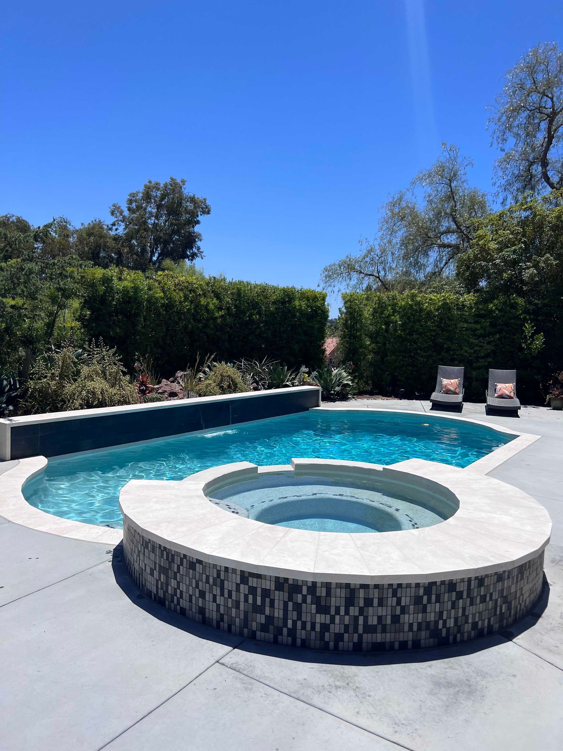 Pool with a built-in spa. Blue water, gray concrete patio, green hedge background, lounge chairs. Sunny day.
