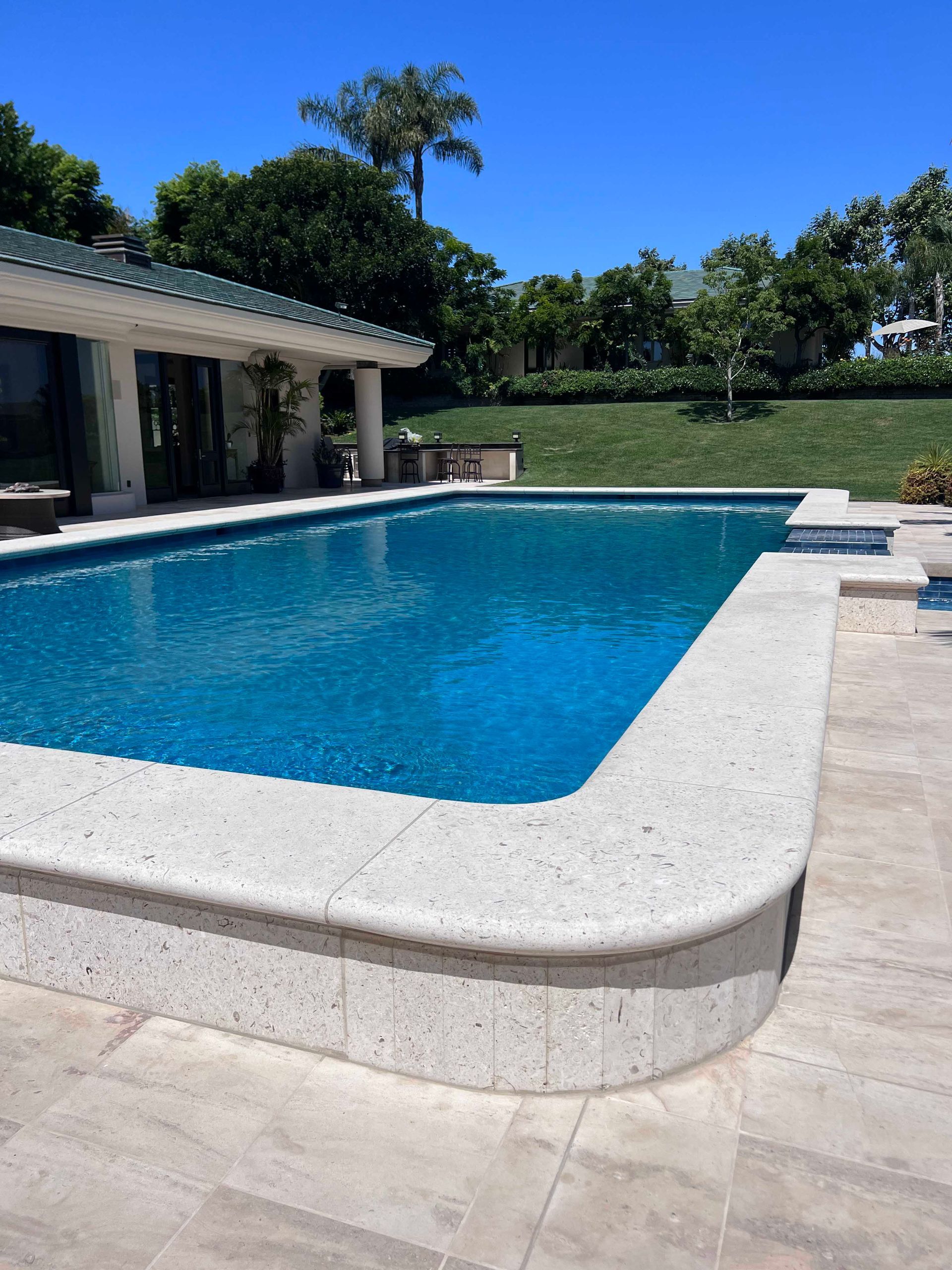 A rectangular swimming pool with turquoise water and a light-colored stone border under a bright blue sky.