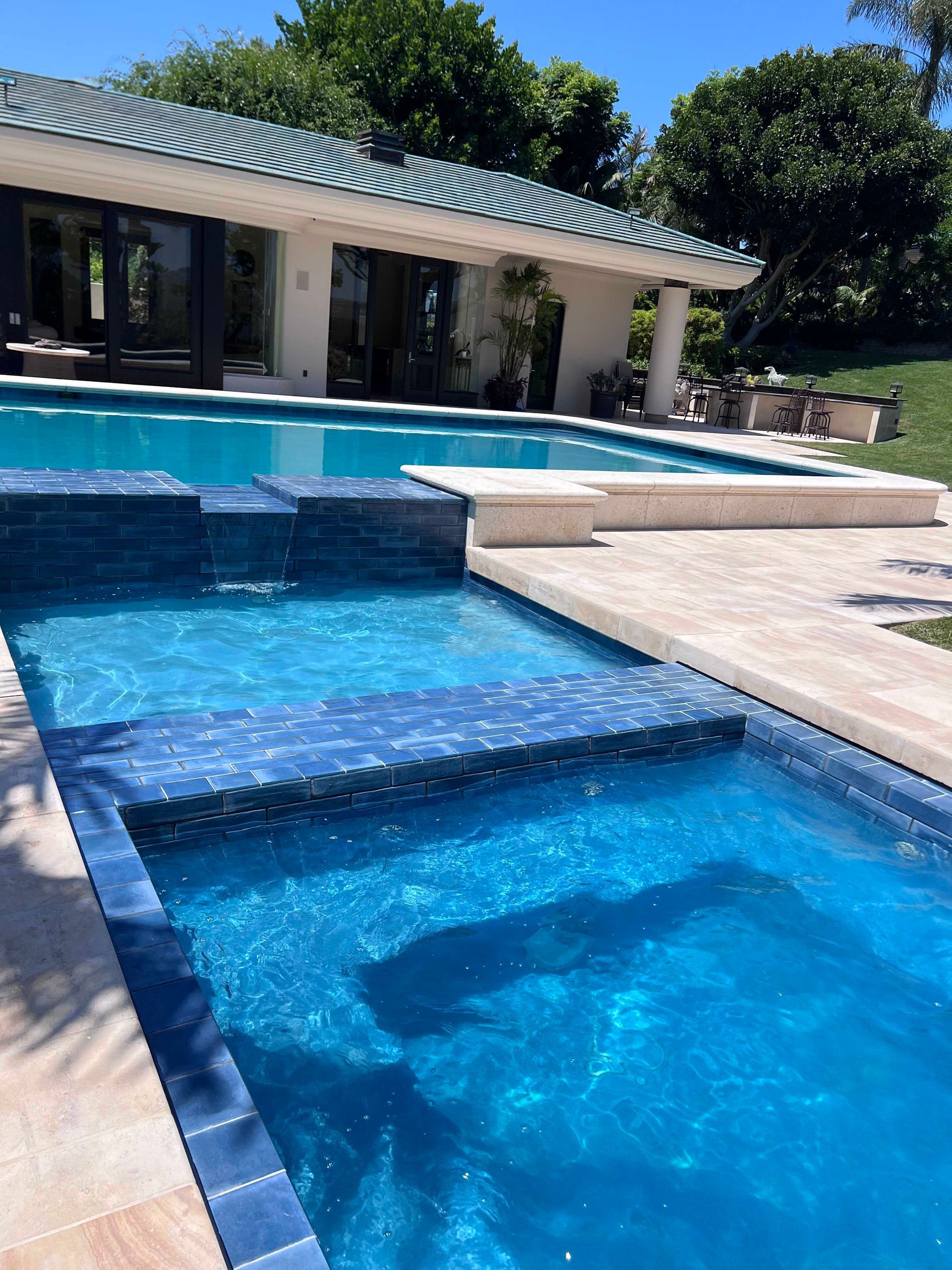 Blue-tiled pool and spa with waterfall feature, adjacent to a patio and building with a green-tiled roof, outdoors on a sunny day.