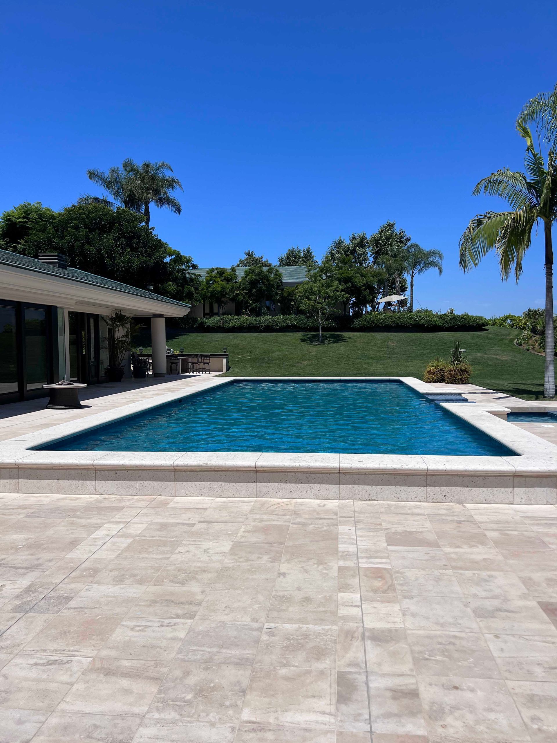 Swimming pool with blue water, surrounded by light-colored stone, next to a house under a clear, blue sky.