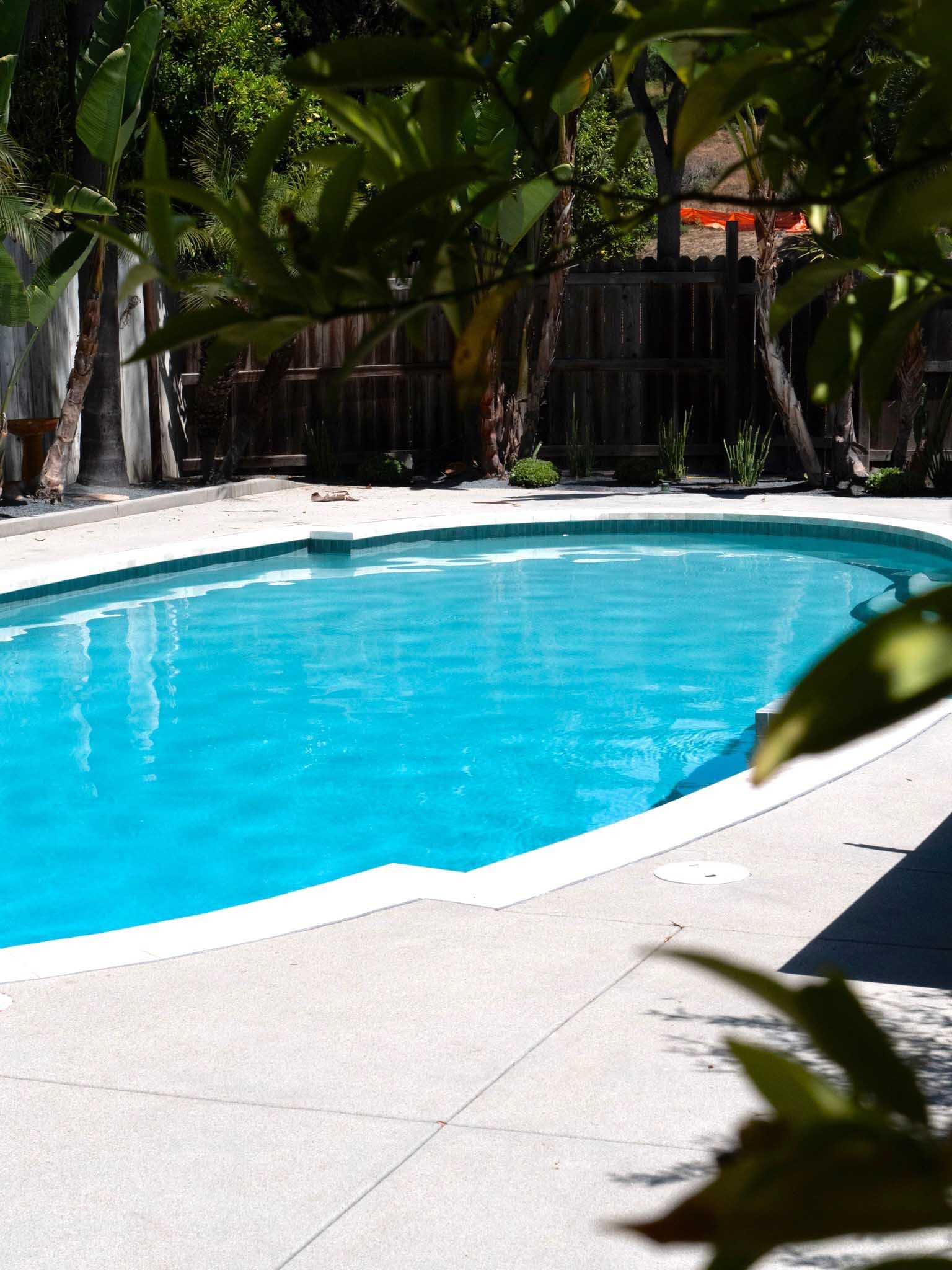 Blue swimming pool surrounded by concrete, with trees and a fence in the background.