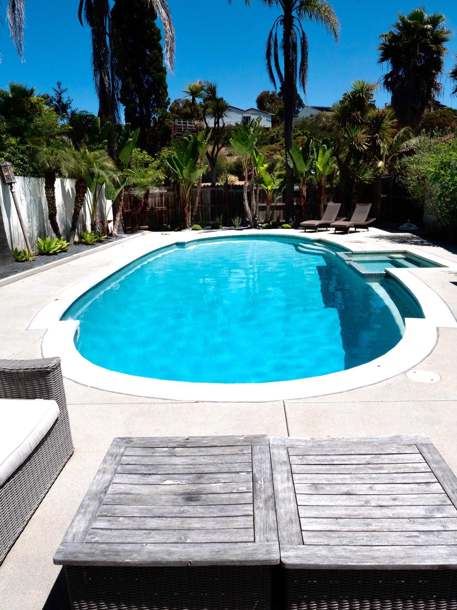 Swimming pool in a sunny backyard with palm trees, lounge chairs, and wooden table.