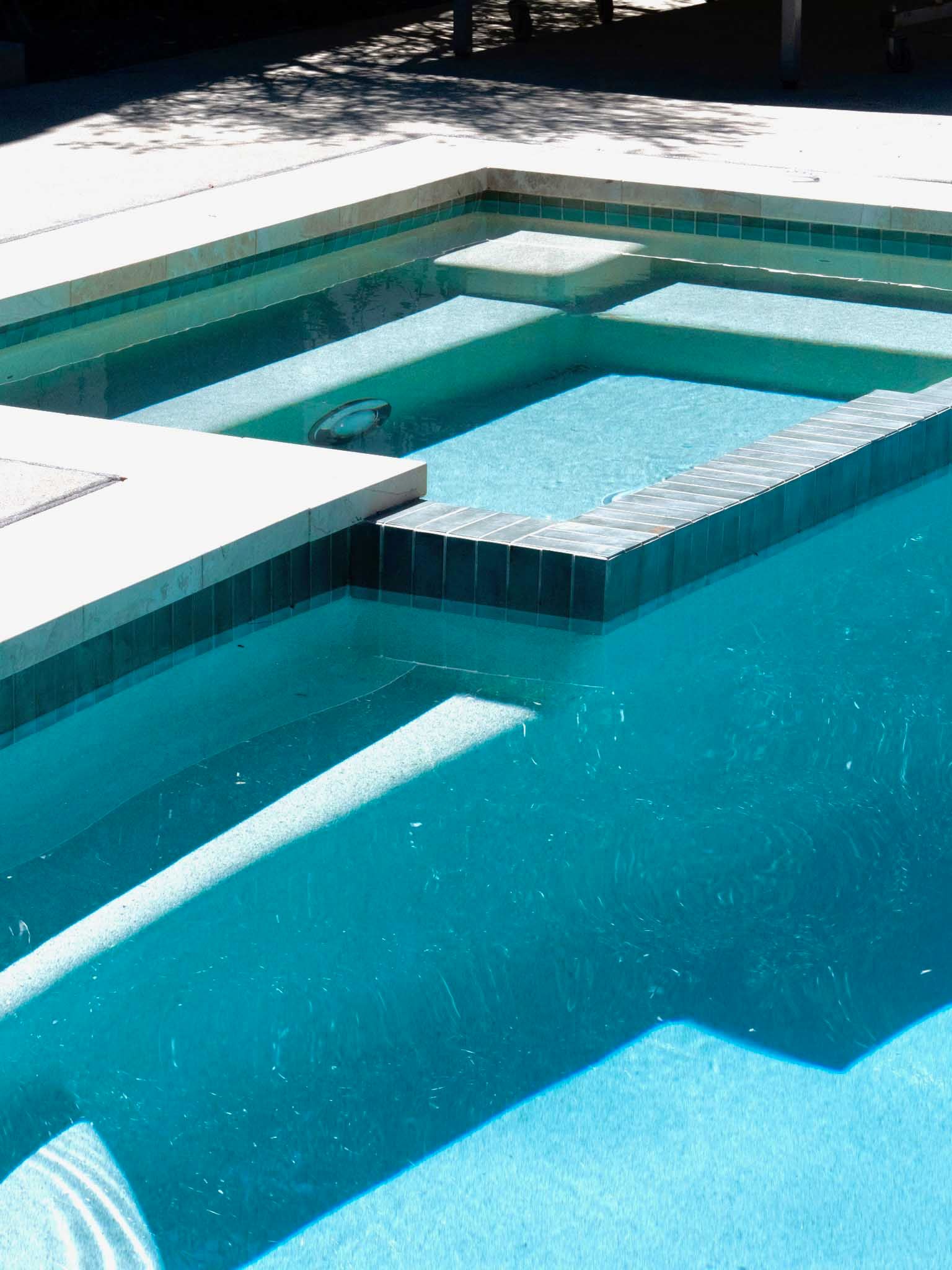 Pool with built-in spa, steps, turquoise water, light-colored concrete coping, and dark tile trim.