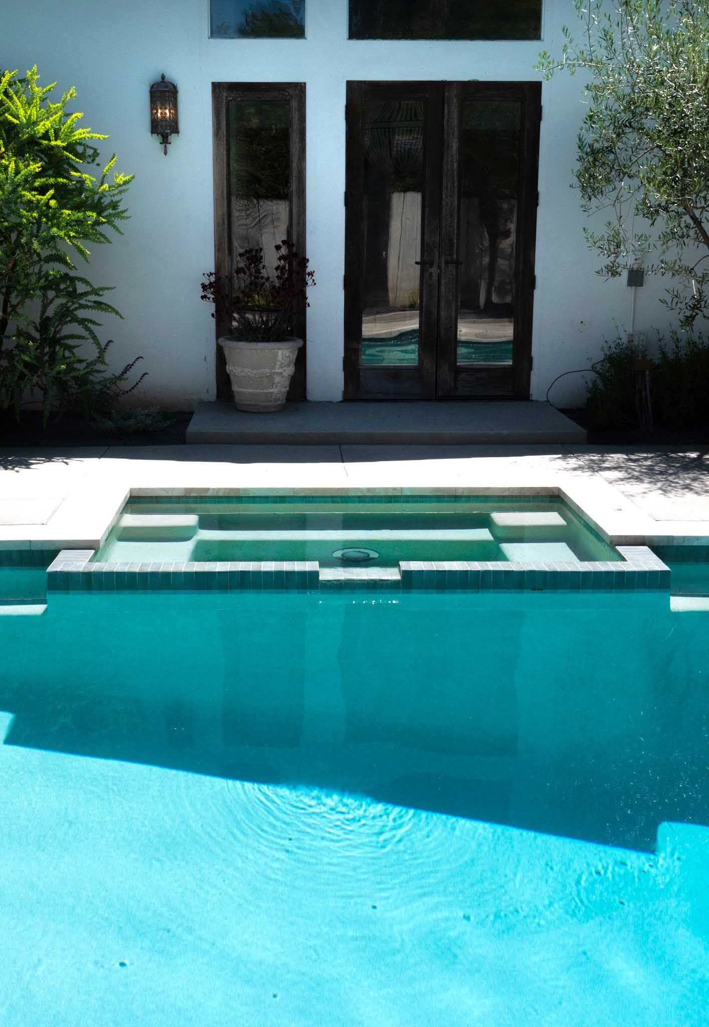 Pool with built-in steps, reflecting sunlight. Dark wood doors and white wall with potted plant behind.