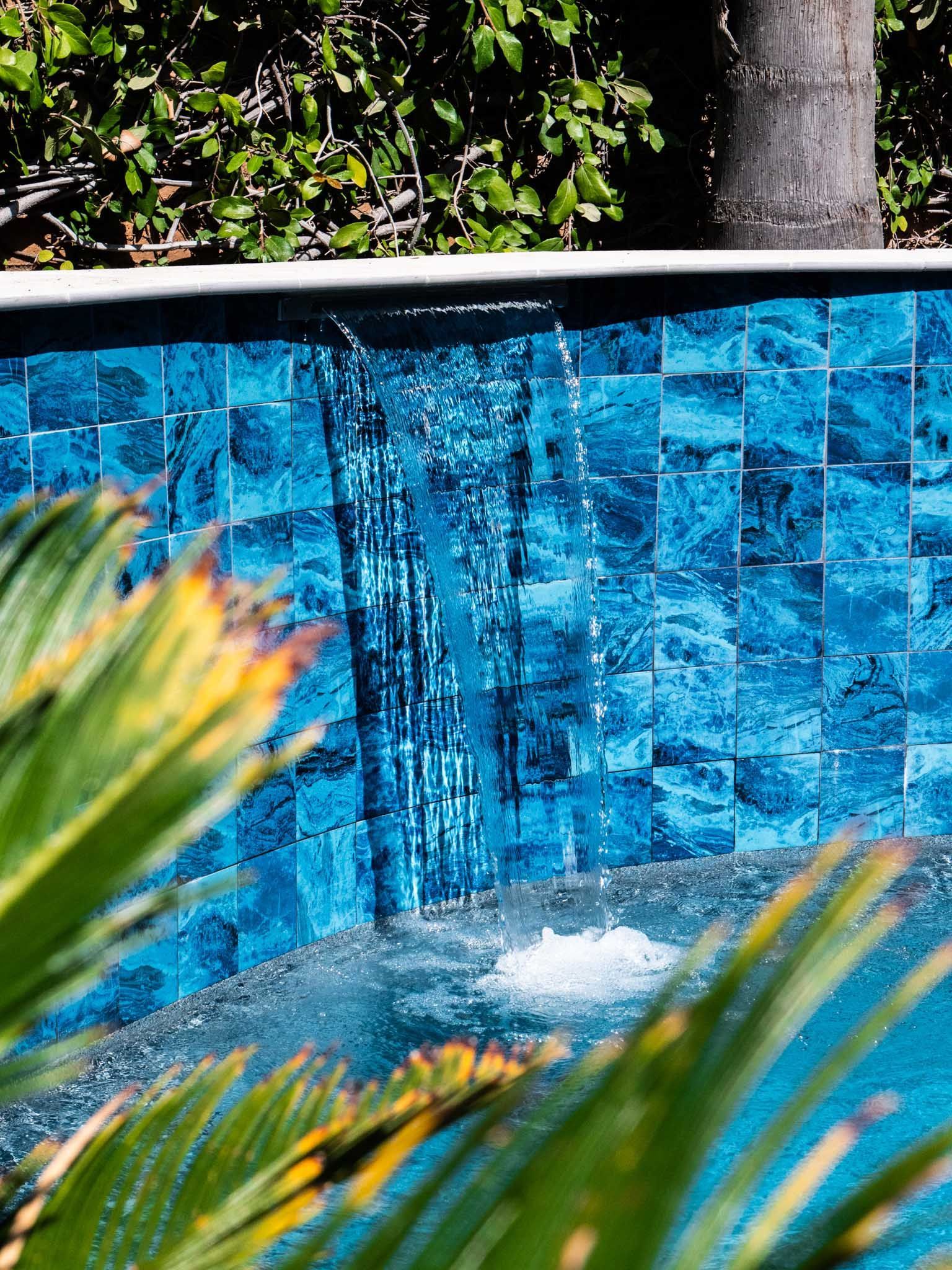 Water cascading down a blue tiled wall into a pool with lush green foliage.