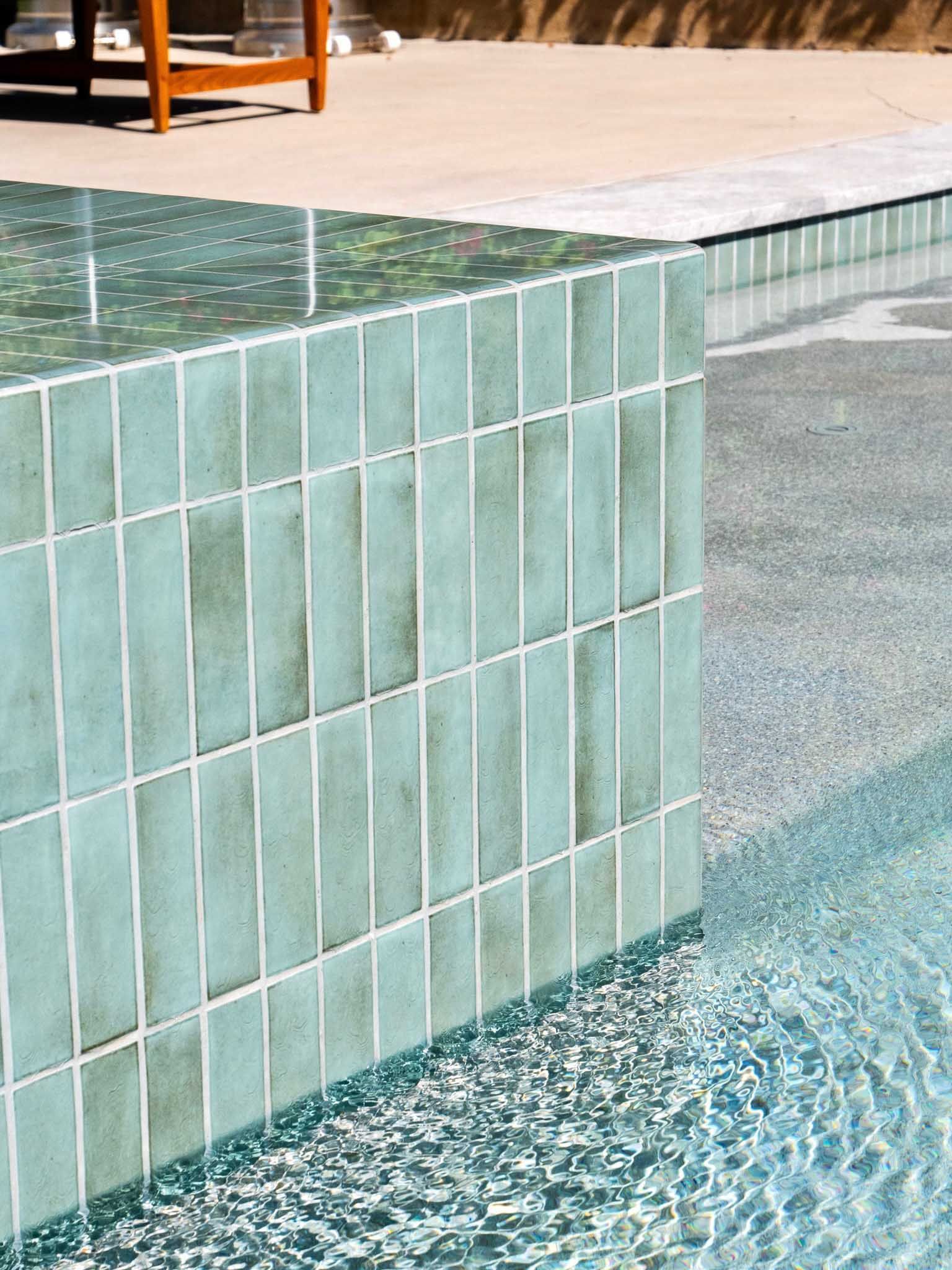 Turquoise tile pool wall with water flowing, concrete patio, and wooden furniture.