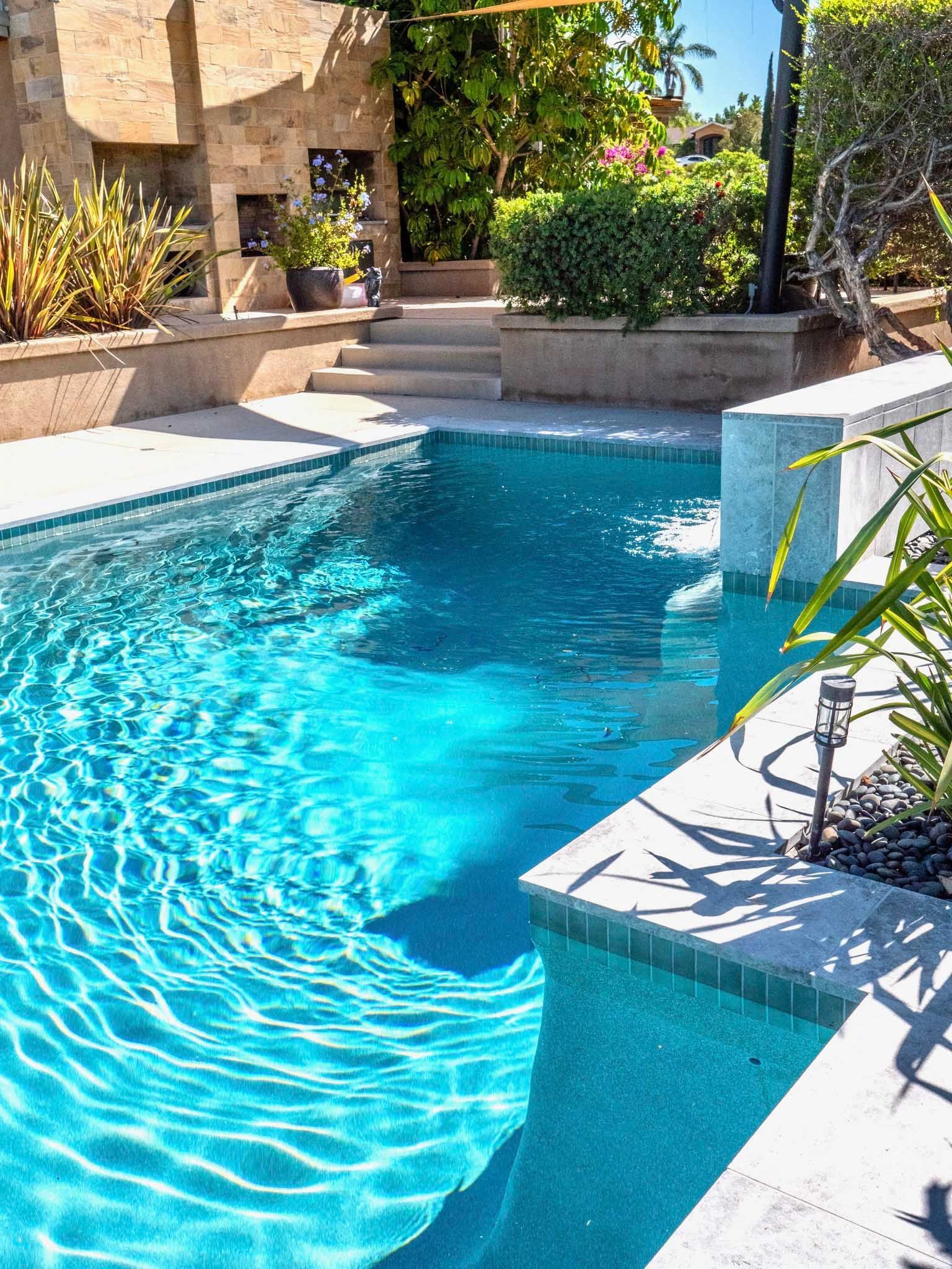 Swimming pool with turquoise water, stone patio, and lush greenery. Sunny day.