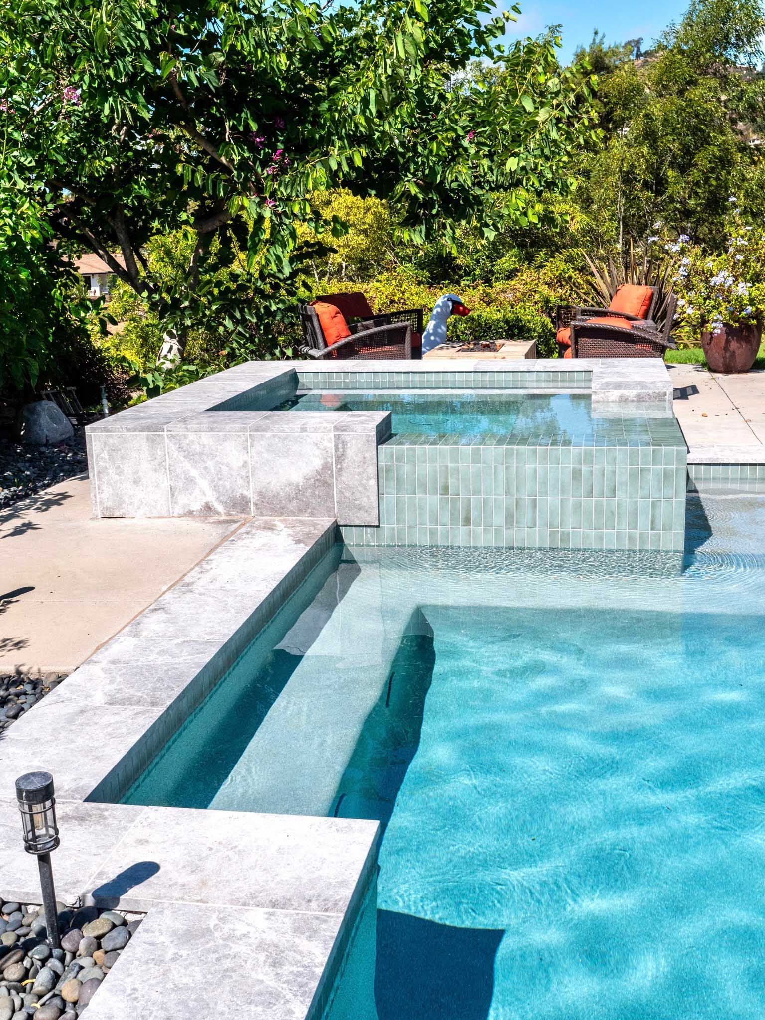 Backyard pool with spa, steps, blue water, stone border. Two chairs, trees in background.