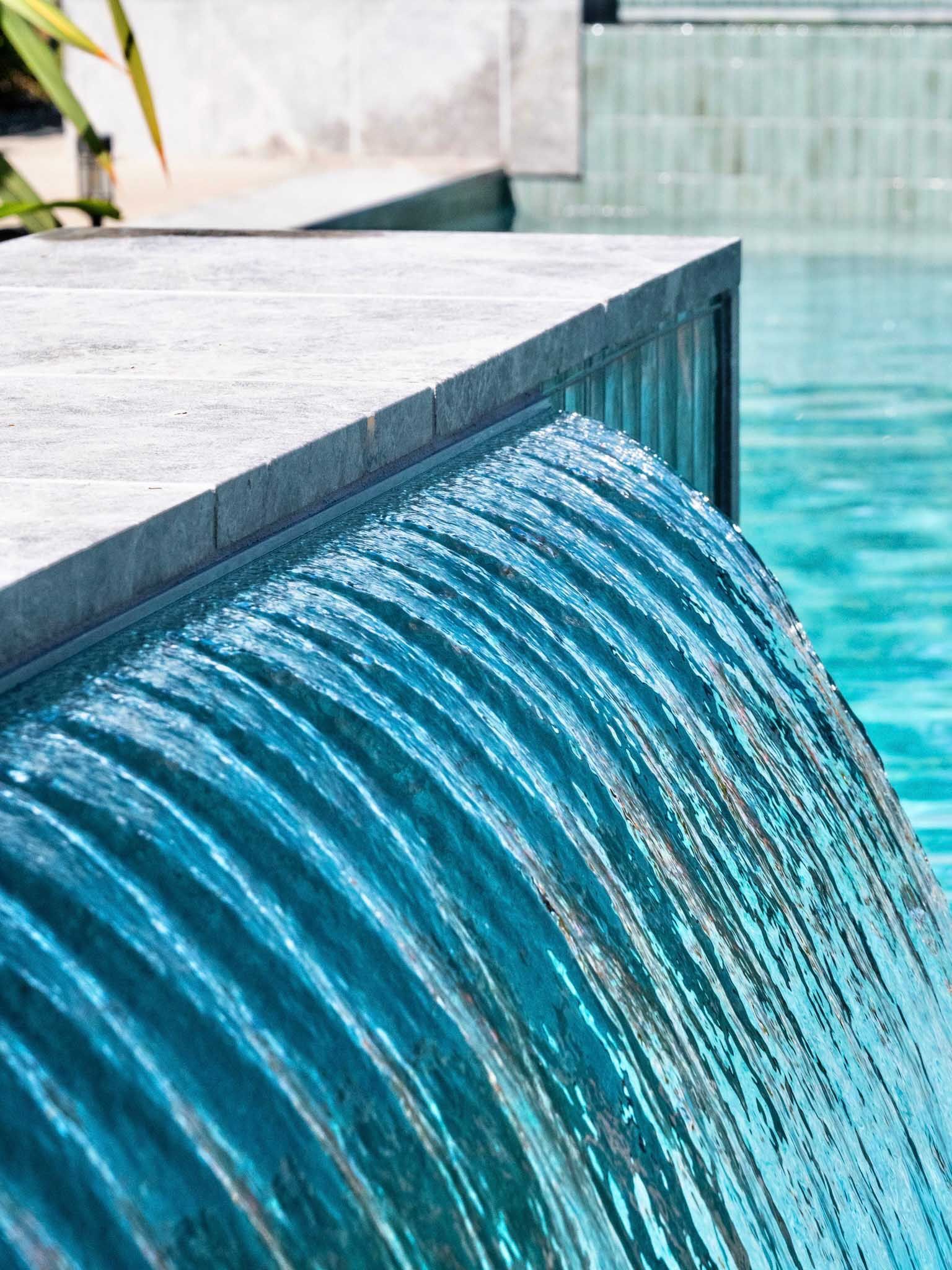 Water flowing over the edge of a turquoise pool into another turquoise pool, a concrete ledge in focus.