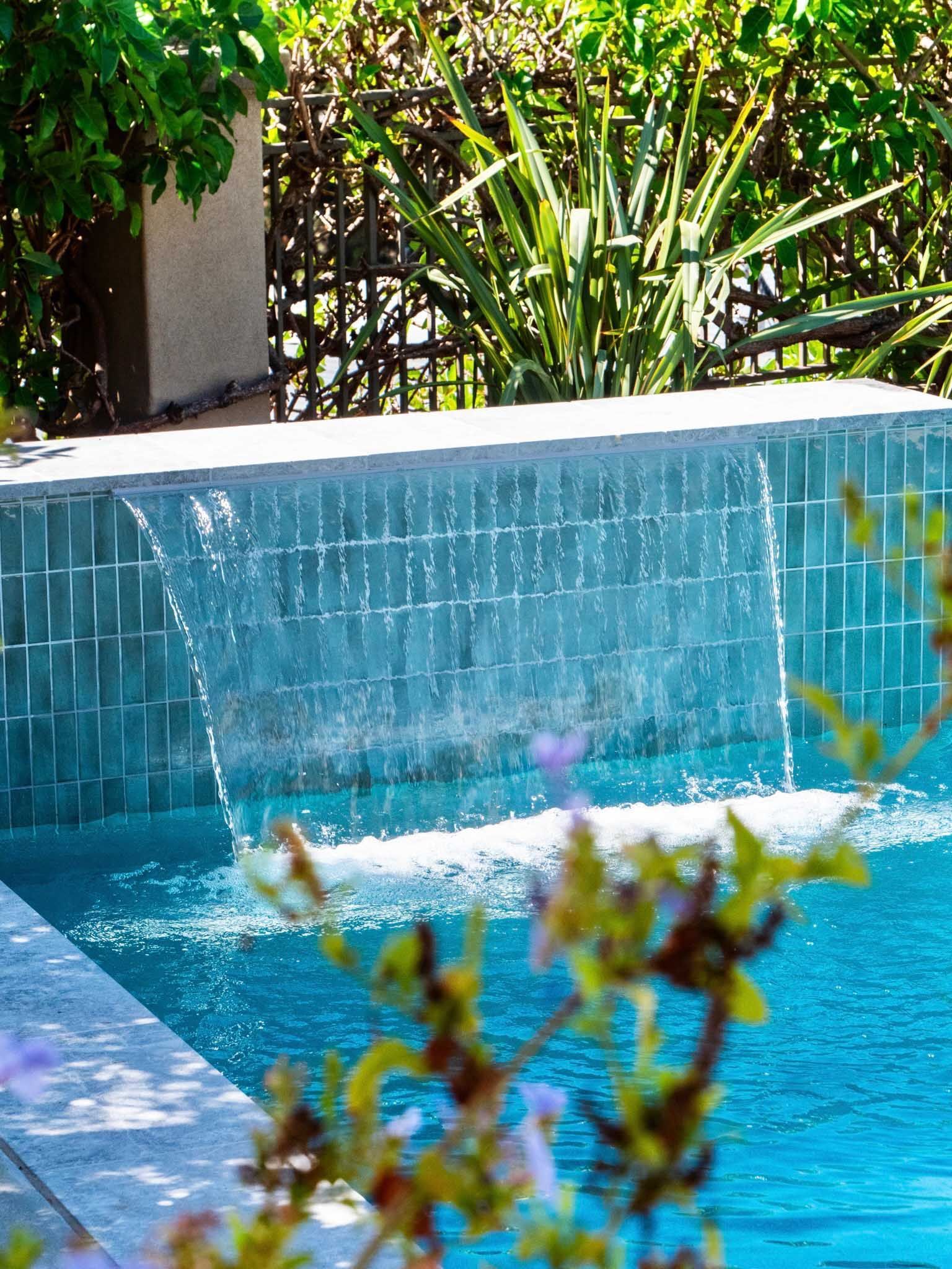 Pool with cascading waterfall, blue tiled wall, and turquoise water. Lush greenery in background.