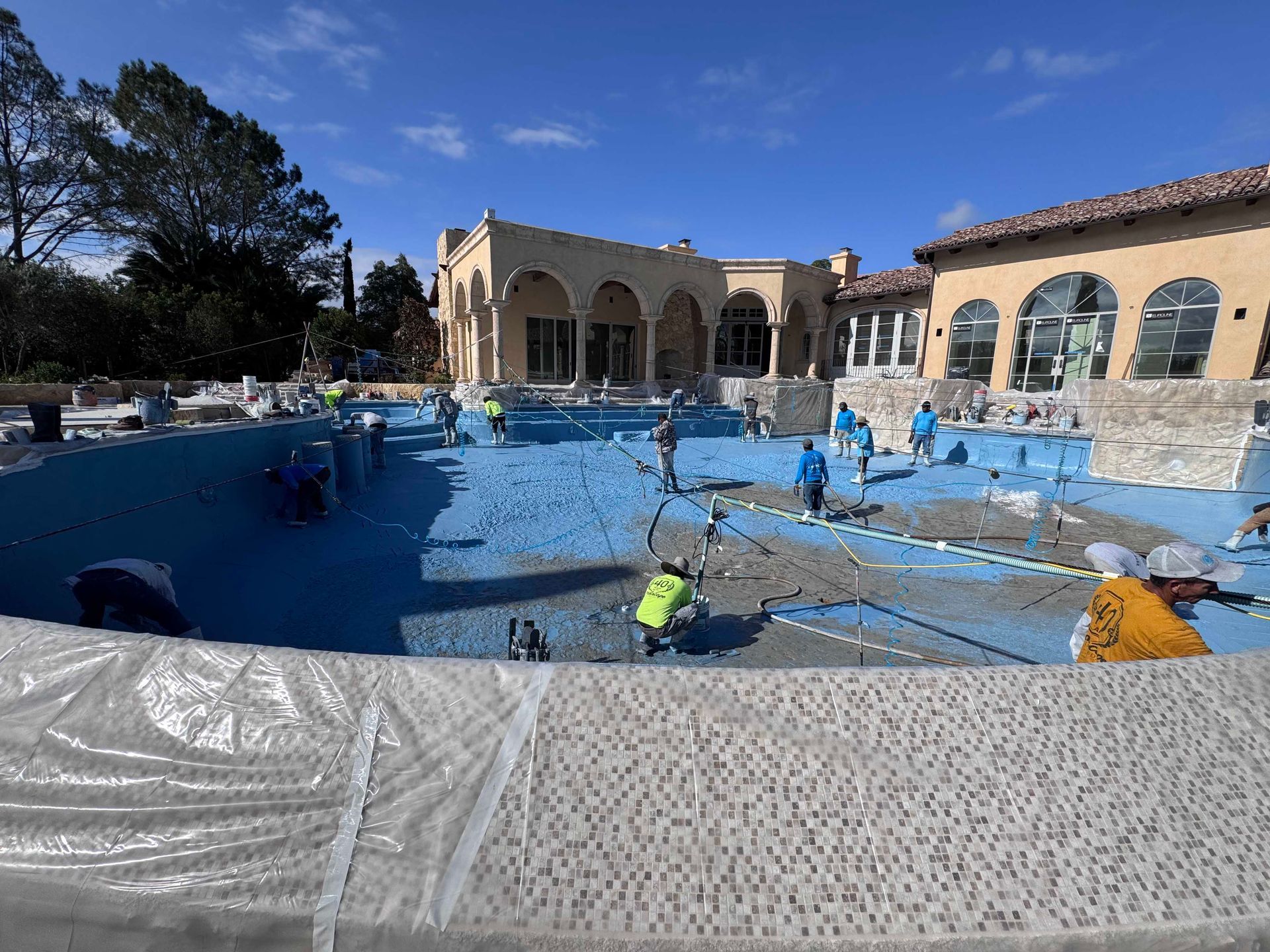 Construction site of a large pool, workers in blue and yellow vests, laying concrete, sunny day.