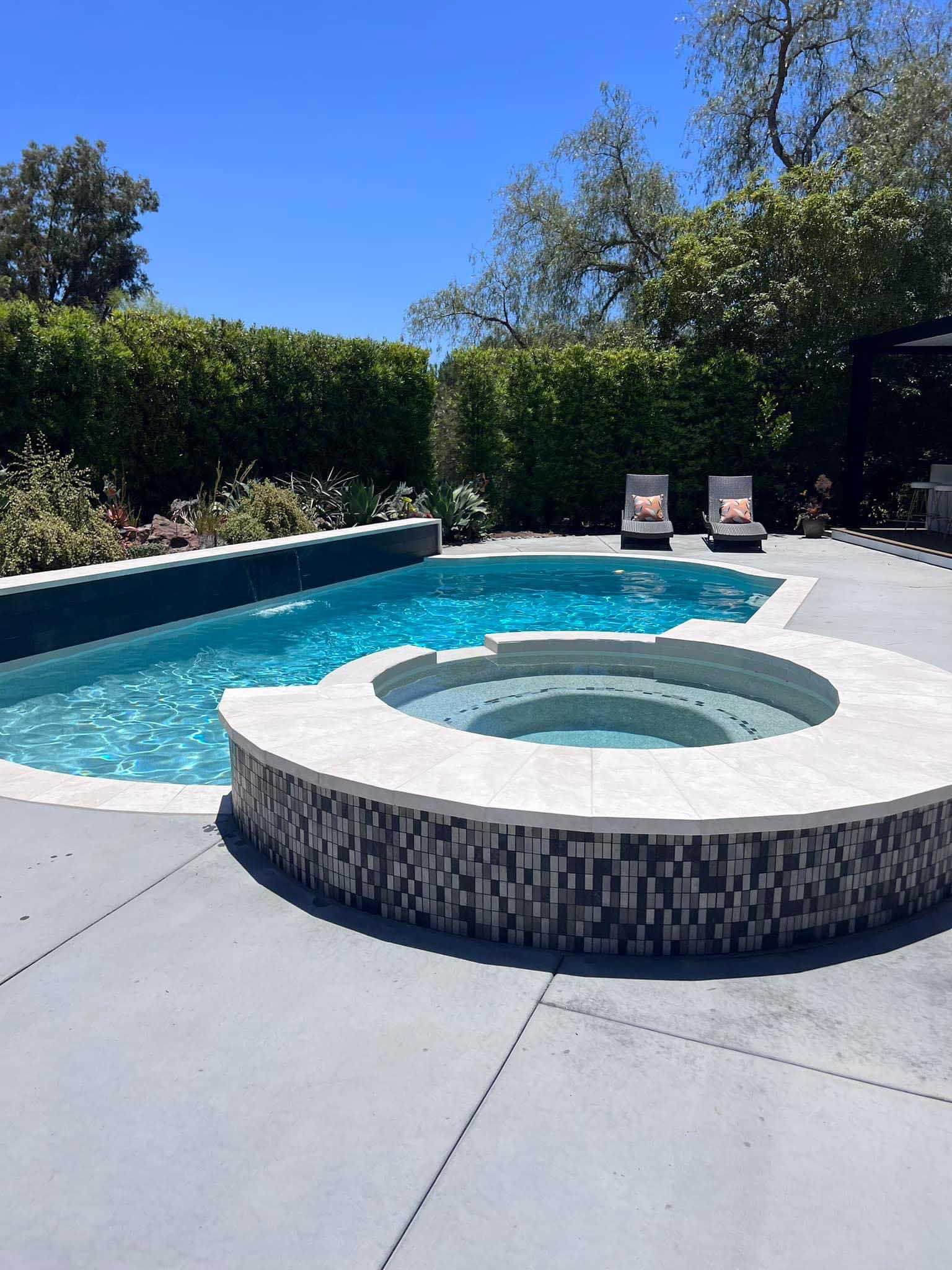 Pool and spa with lounge chairs on a sunny patio, surrounded by hedges and trees.