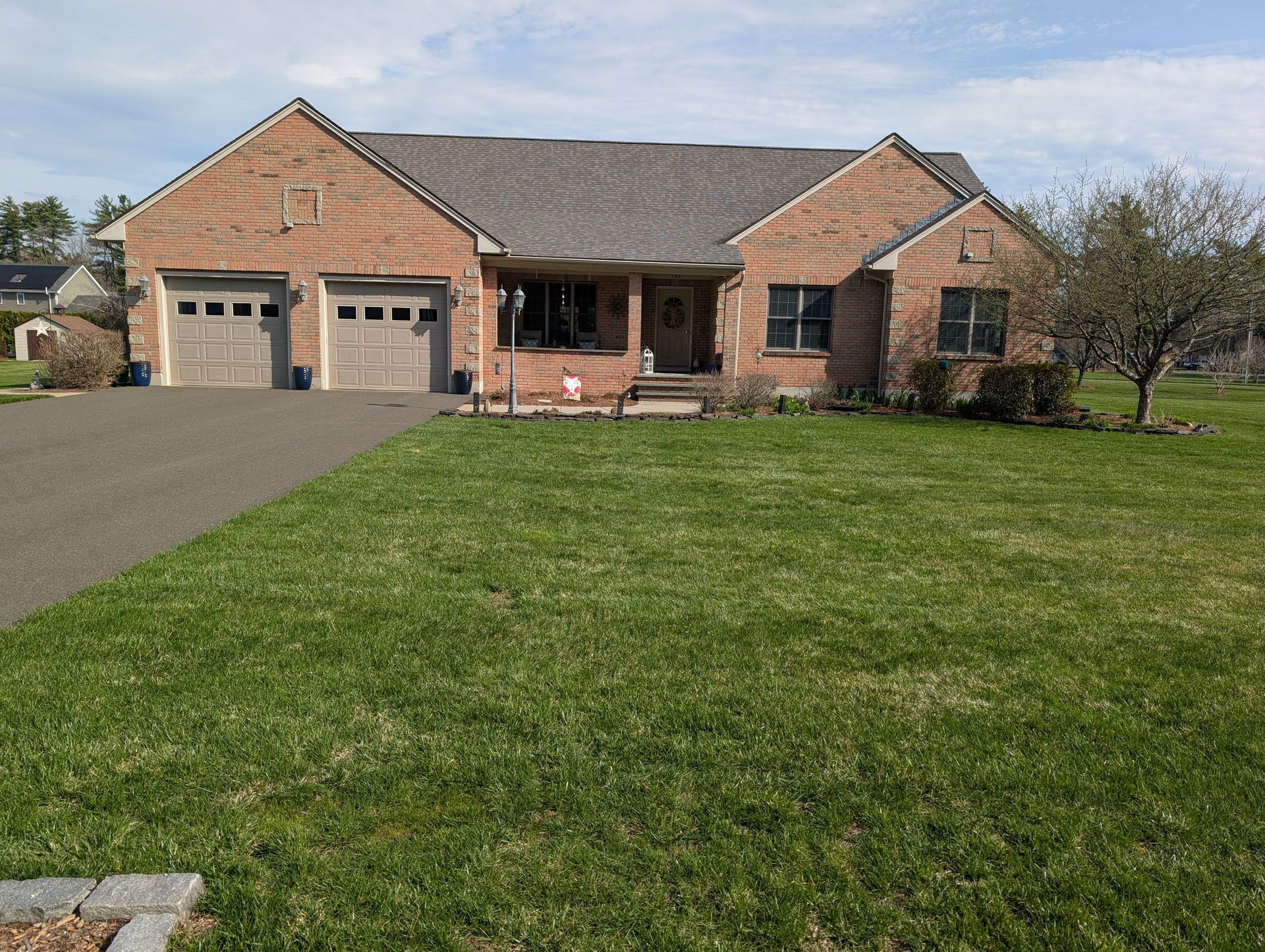 A tan brick, single-story house with a two-car garage, gray roof, and a large front lawn under a blue sky.