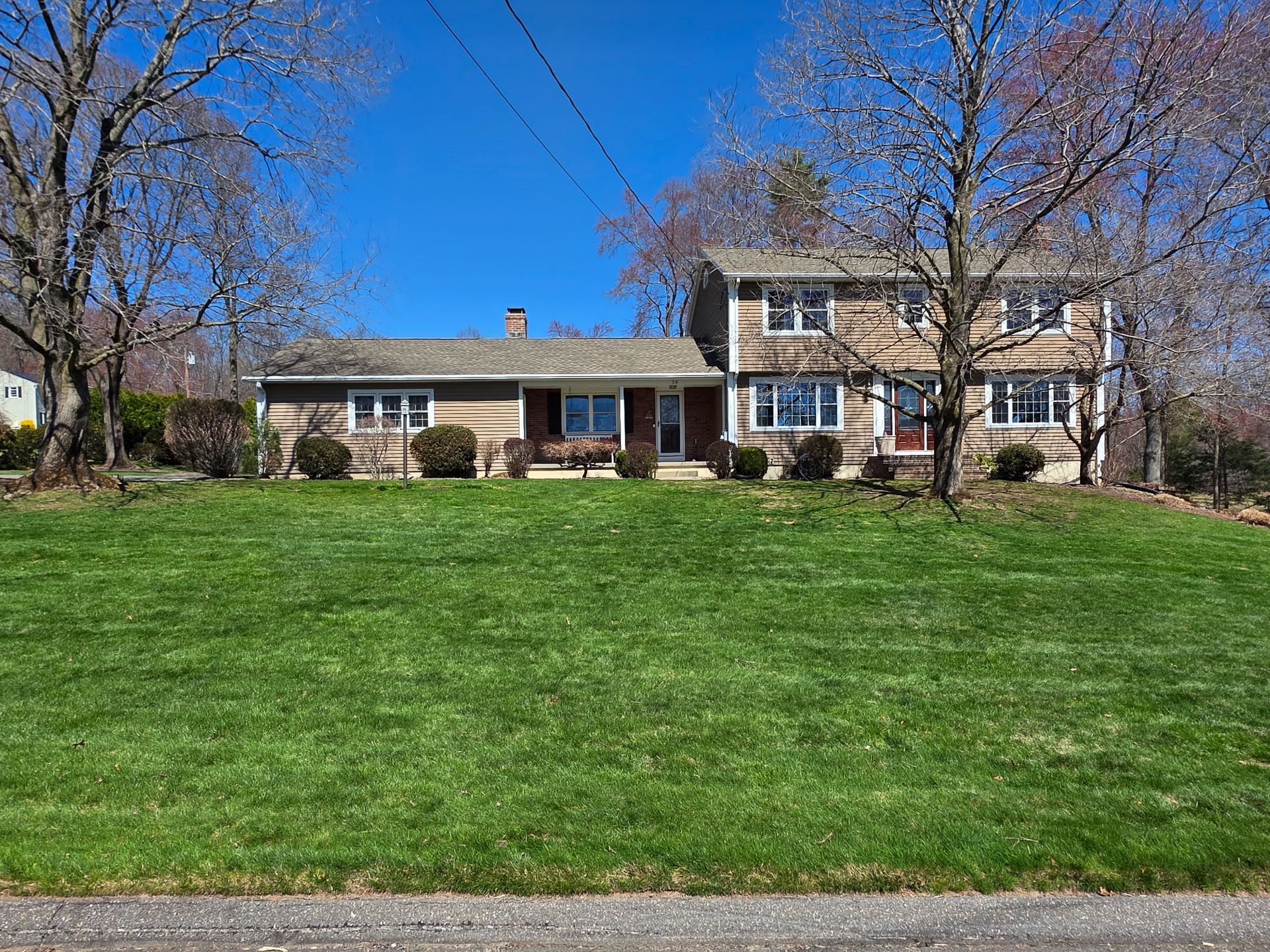 A two-story, tan, shingled suburban house with a green lawn and bare trees under a bright blue sky.