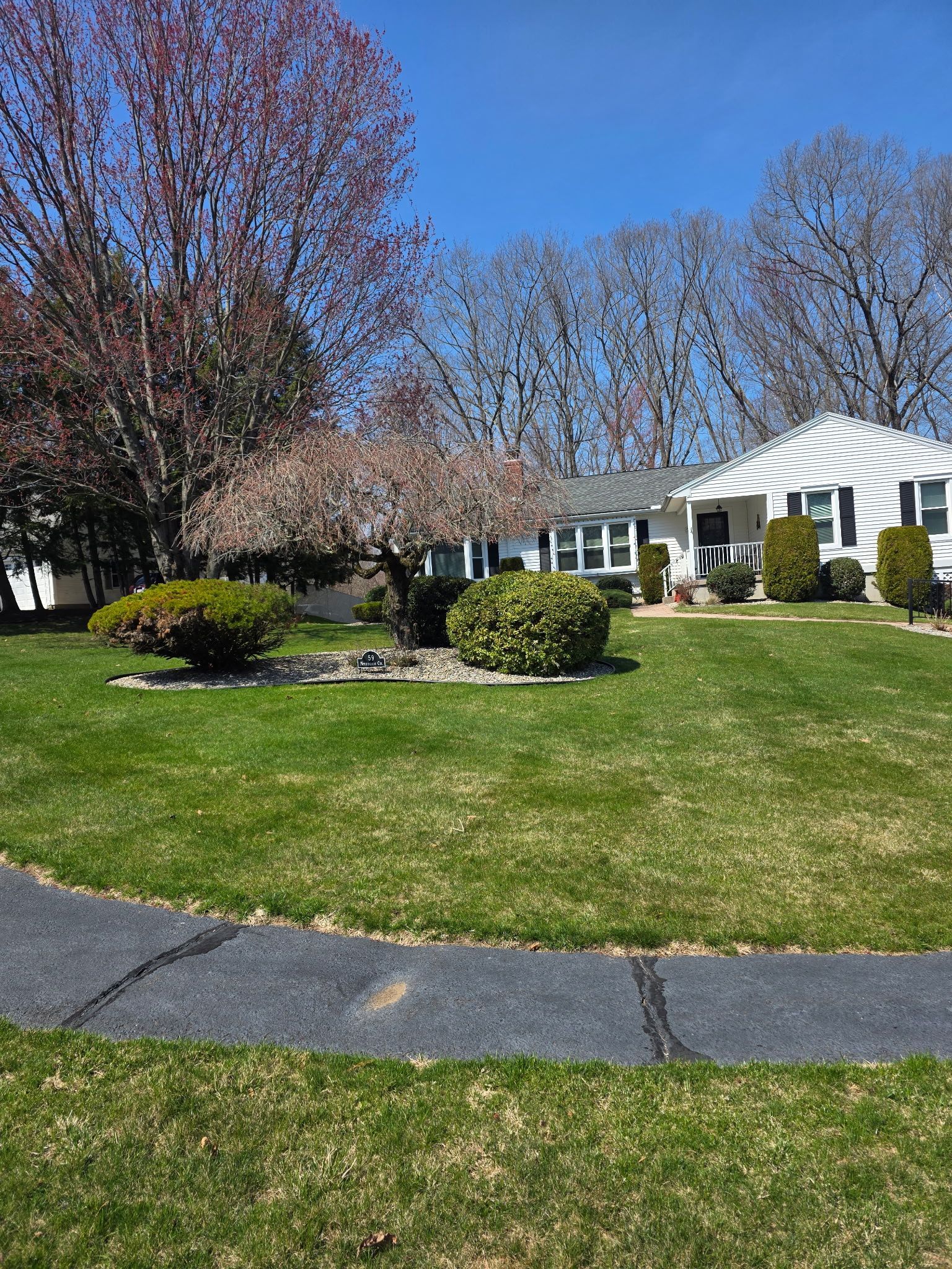 A white house with black shutters sits on a grassy lot with trimmed bushes and trees under a clear blue sky.