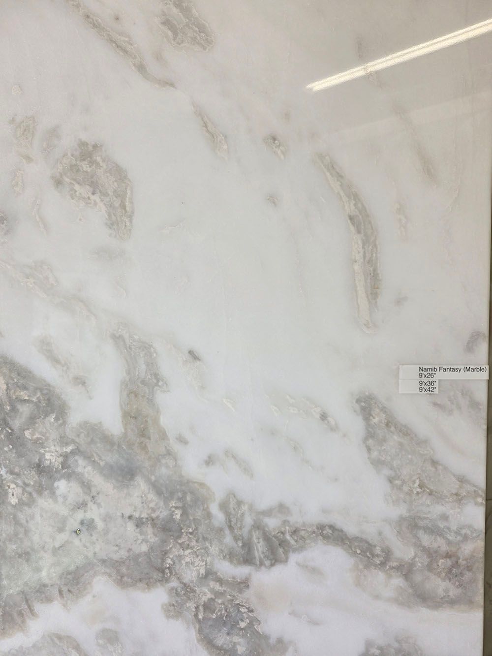 A polished marble slab featuring a white background with swirling, soft grey and cream veining.