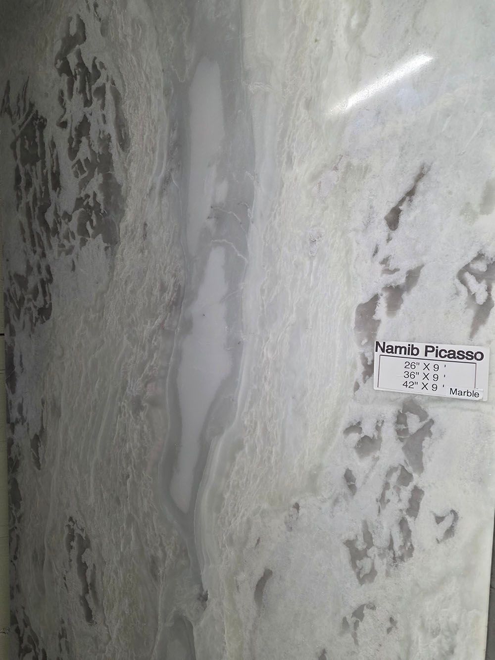 A large, polished slab of white and grey Namib Picasso marble featuring swirling, fluid patterns and a small label.
