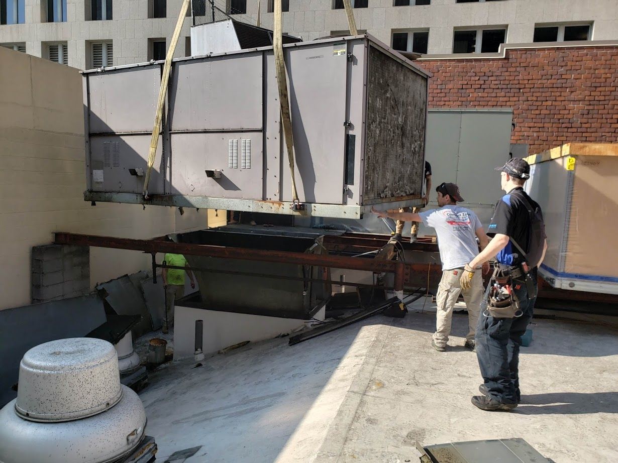HVAC unit being lowered onto a rooftop with two workers assisting. Beige building exterior and clear sky.