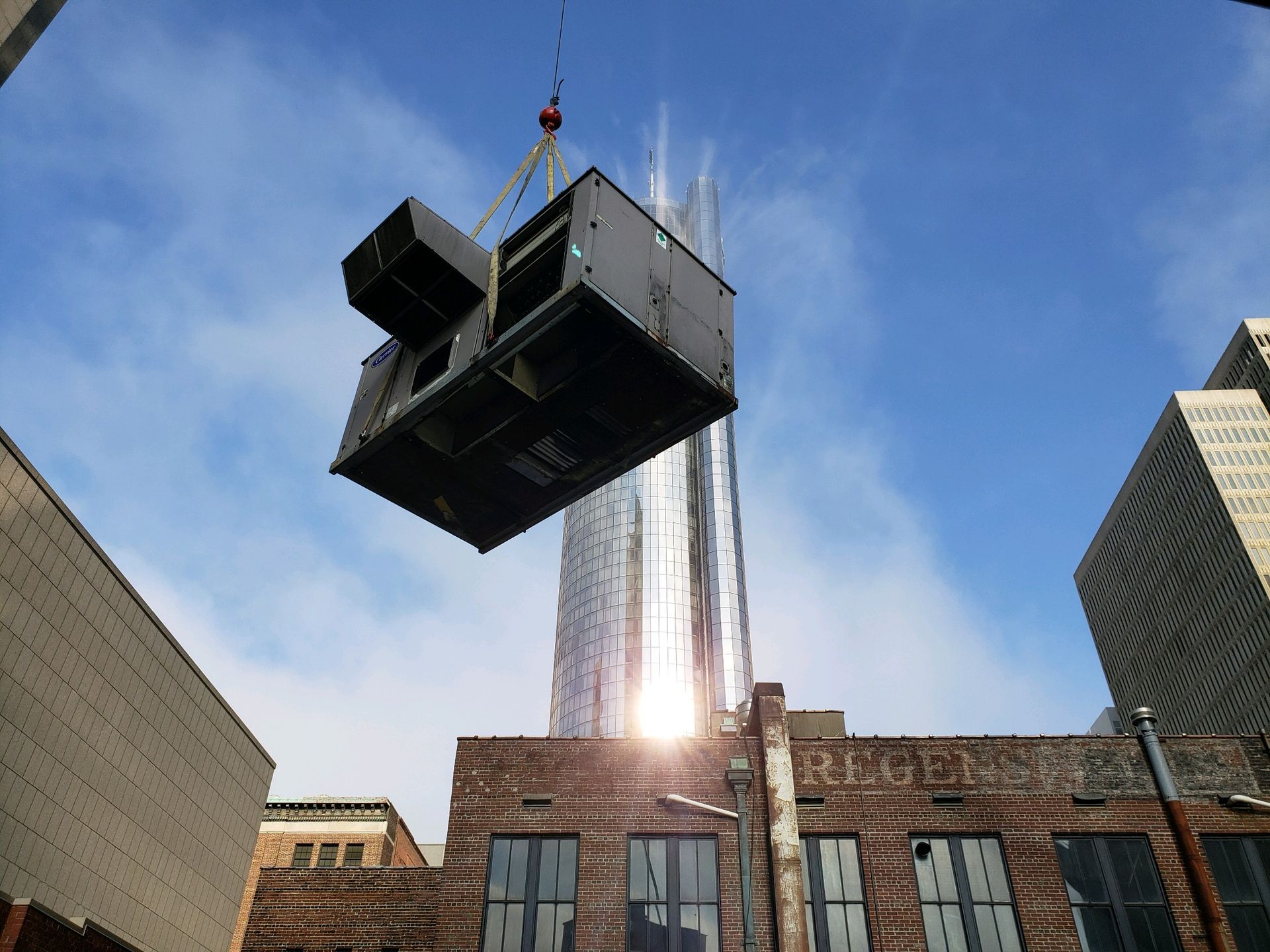 Crane lifting HVAC unit over brick building and tall skyscraper on sunny day.