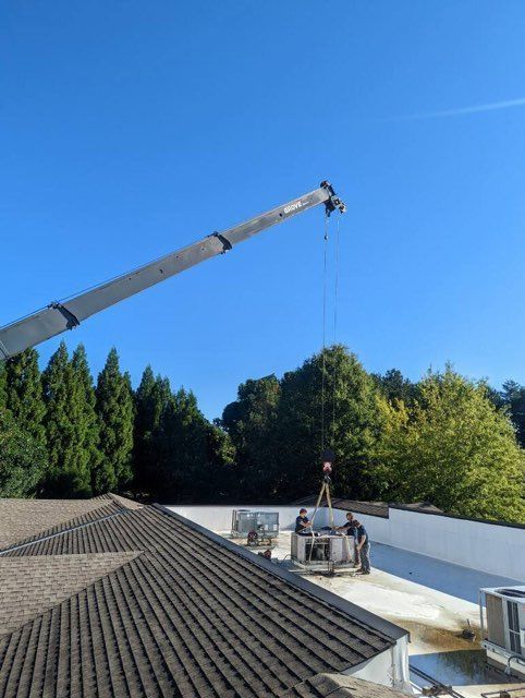 Crane lifting an HVAC unit onto a building roof. Blue sky, green trees, workers present.