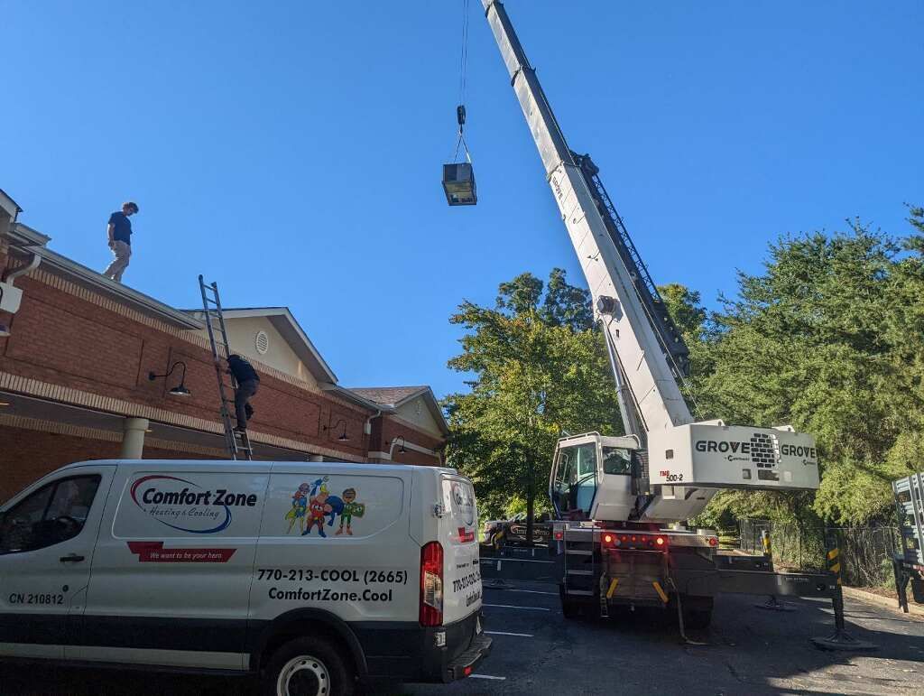 Crane lifting HVAC unit onto a building's roof with workers present. White Comfort Zone van is in front.