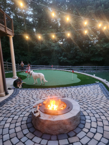 A backyard with a fire pit, putting green, and string lights; people and dogs are present.