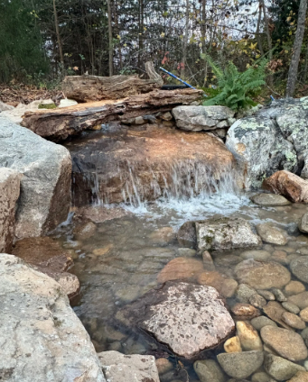 Small waterfall cascading over rocks into a clear, shallow stream with rounded stones.