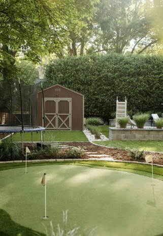 Backyard with a brown shed, putting green, trampoline, and lush landscaping.