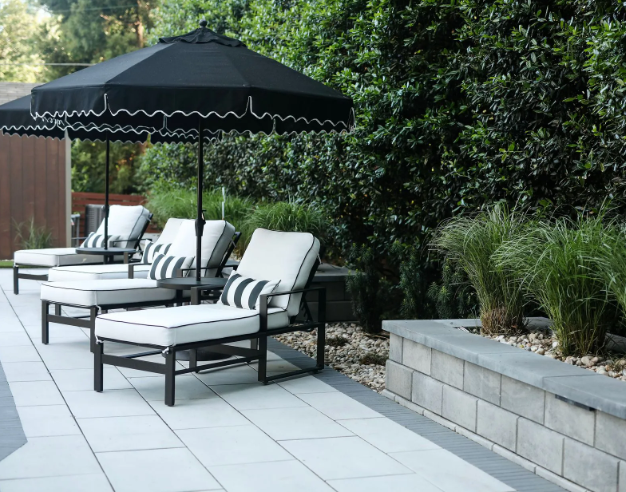 Lounge chairs with striped cushions under black umbrellas on a patio. Green shrubs border the area.