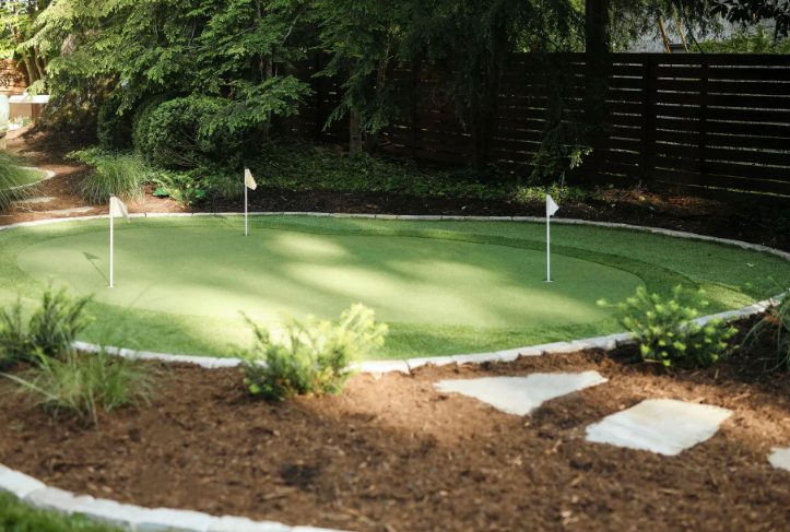 Artificial green golf putting area with three flags, surrounded by mulch, stones, and landscaping.