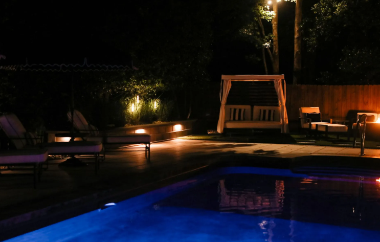 Nighttime outdoor scene with pool, lit cabana, lounge chairs, and string lights.