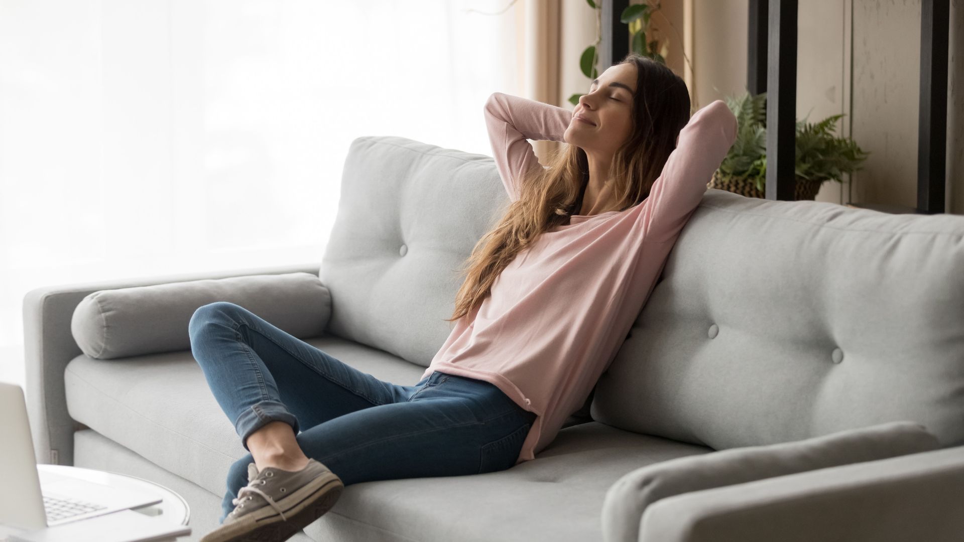 Woman relaxing on a gray sofa with arms behind her head, eyes closed. Sunlight, light pink top, jeans.