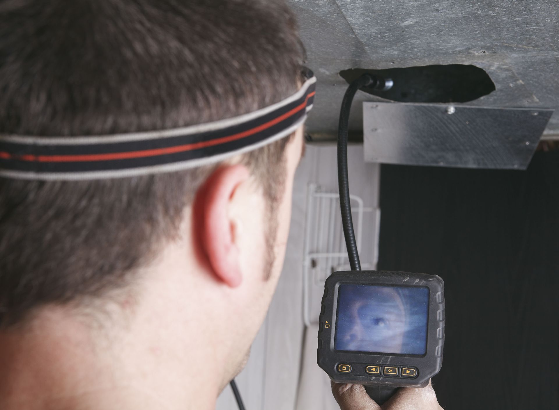 Man using a borescope to inspect a hole in ductwork. He wears a headlamp and holds a screen.