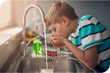 A young boy drinks water from a faucet in a kitchen sink.