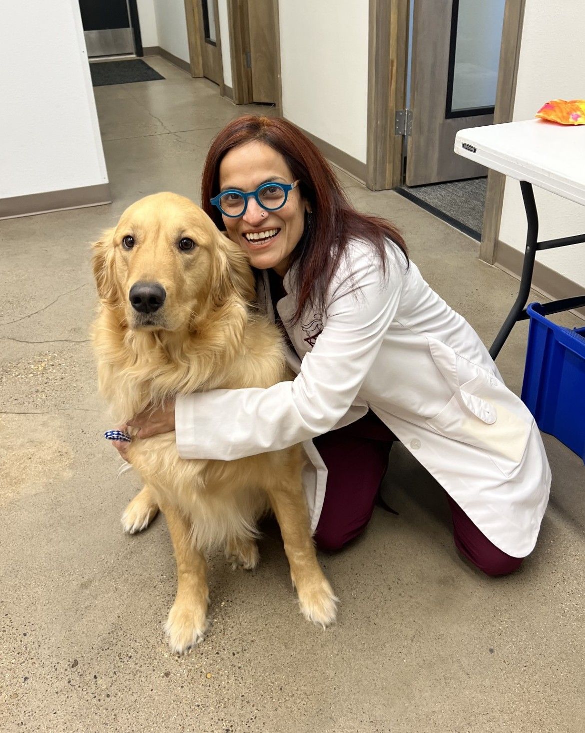 Woman in lab coat hugs a Golden Retriever. Both are smiling. Indoors, near a table and door.