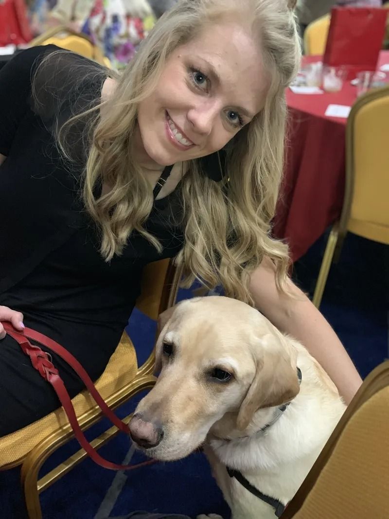 Woman in black dress smiles, petting a yellow Labrador dog sitting near her.