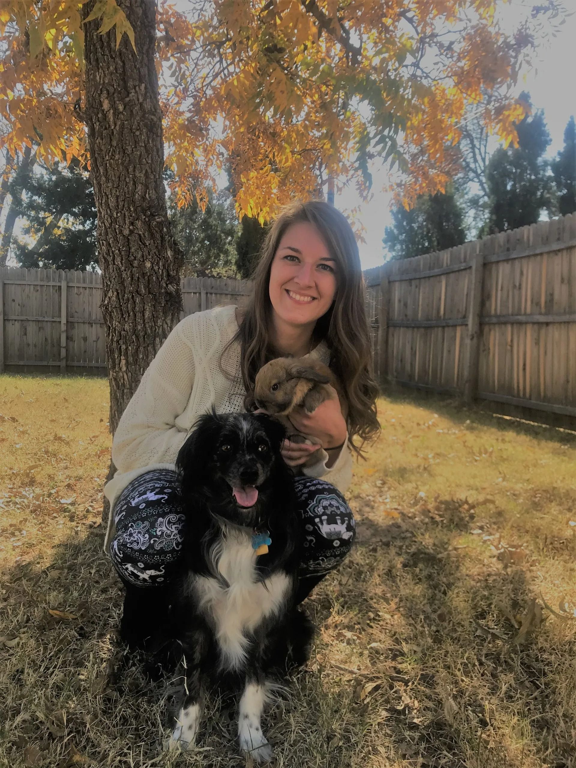 Woman smiles outdoors, holding two small pets, a dog and a hamster, with fall foliage behind.