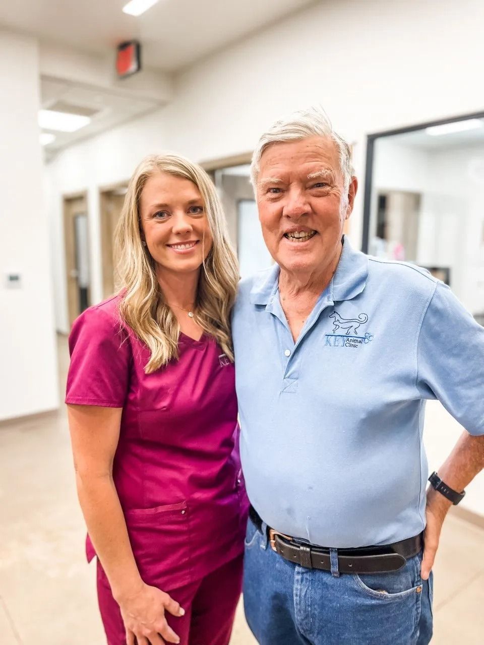 Woman in pink scrubs and older man in blue shirt posing in a hallway, smiling.