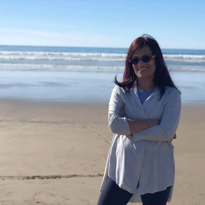Woman with sunglasses and arms crossed at the beach, wearing a light gray sweater. Blue sky and ocean background.