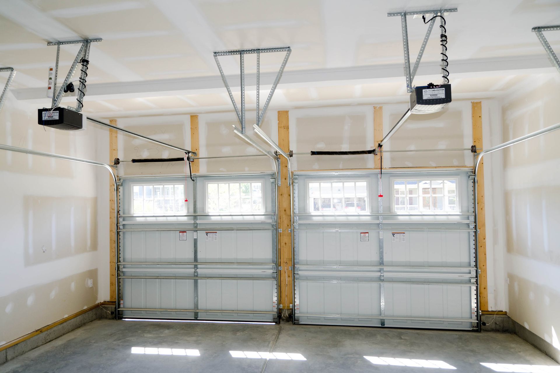Inside a newly constructed garage, two closed white garage doors with window panels. Electric door openers are mounted overhead.