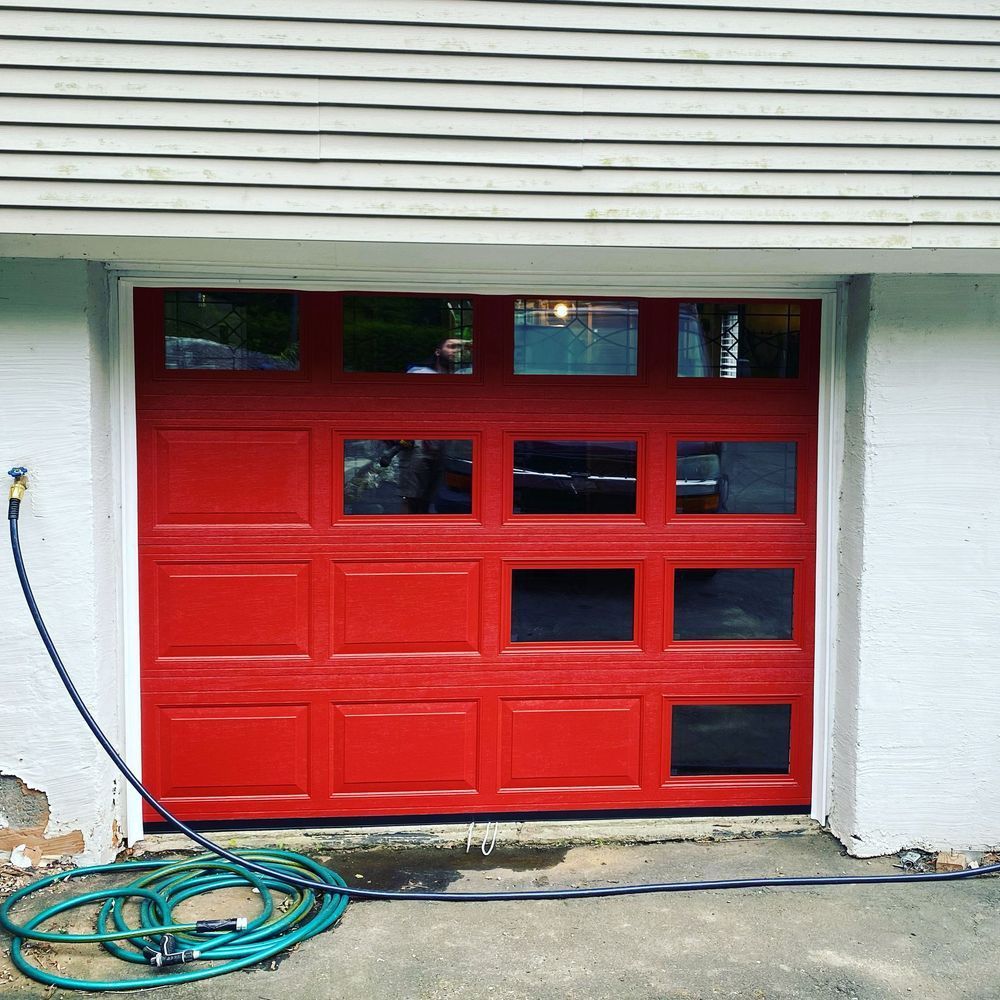 Red garage door with window panels, white trim, and a coiled blue garden hose on concrete.