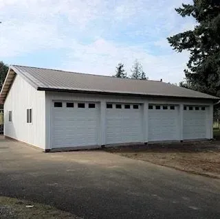 White three-car garage with a sloped roof beside a driveway, surrounded by trees.